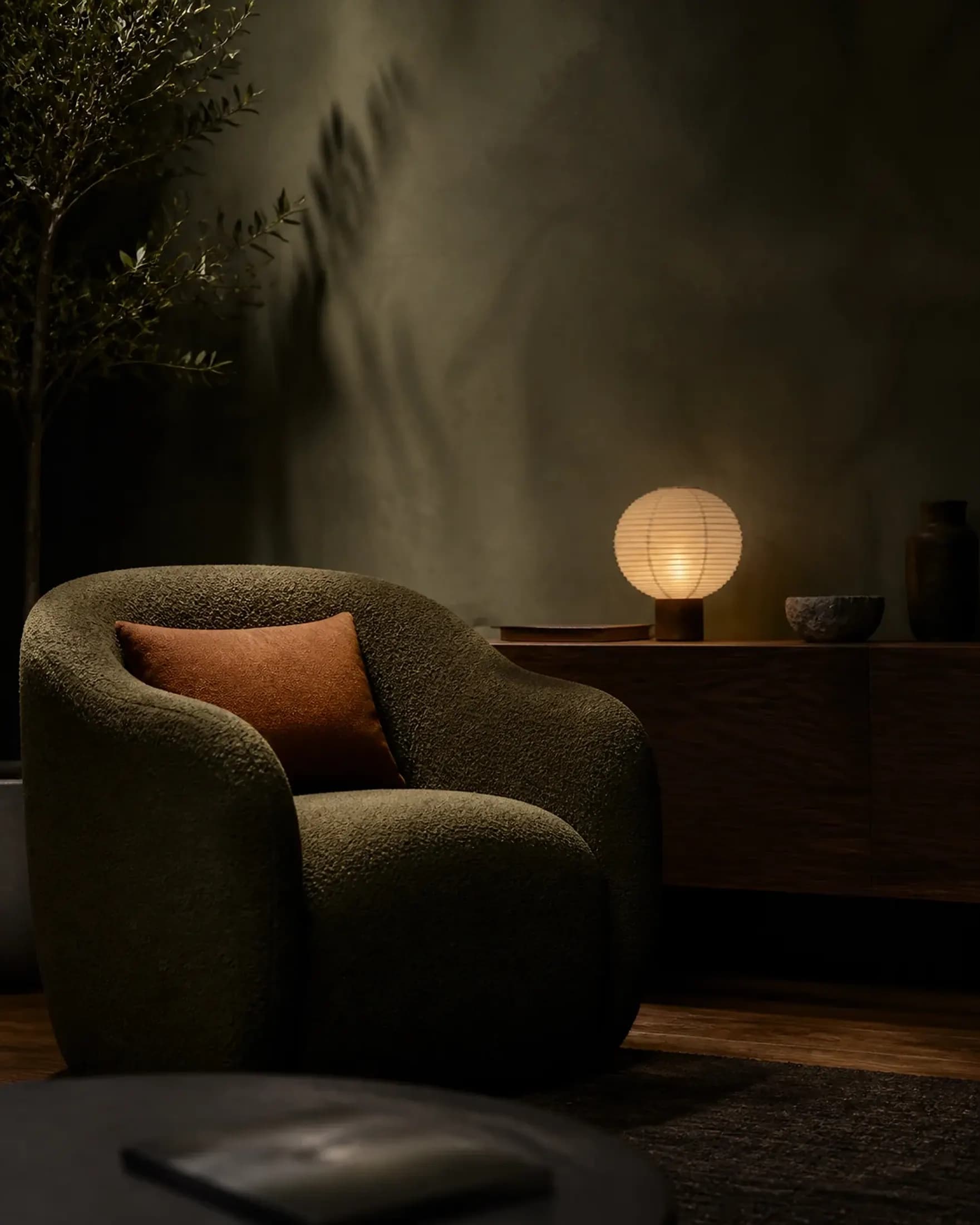Concept image of an olive boucle armchair and rust cushion beside a warm paper lamp