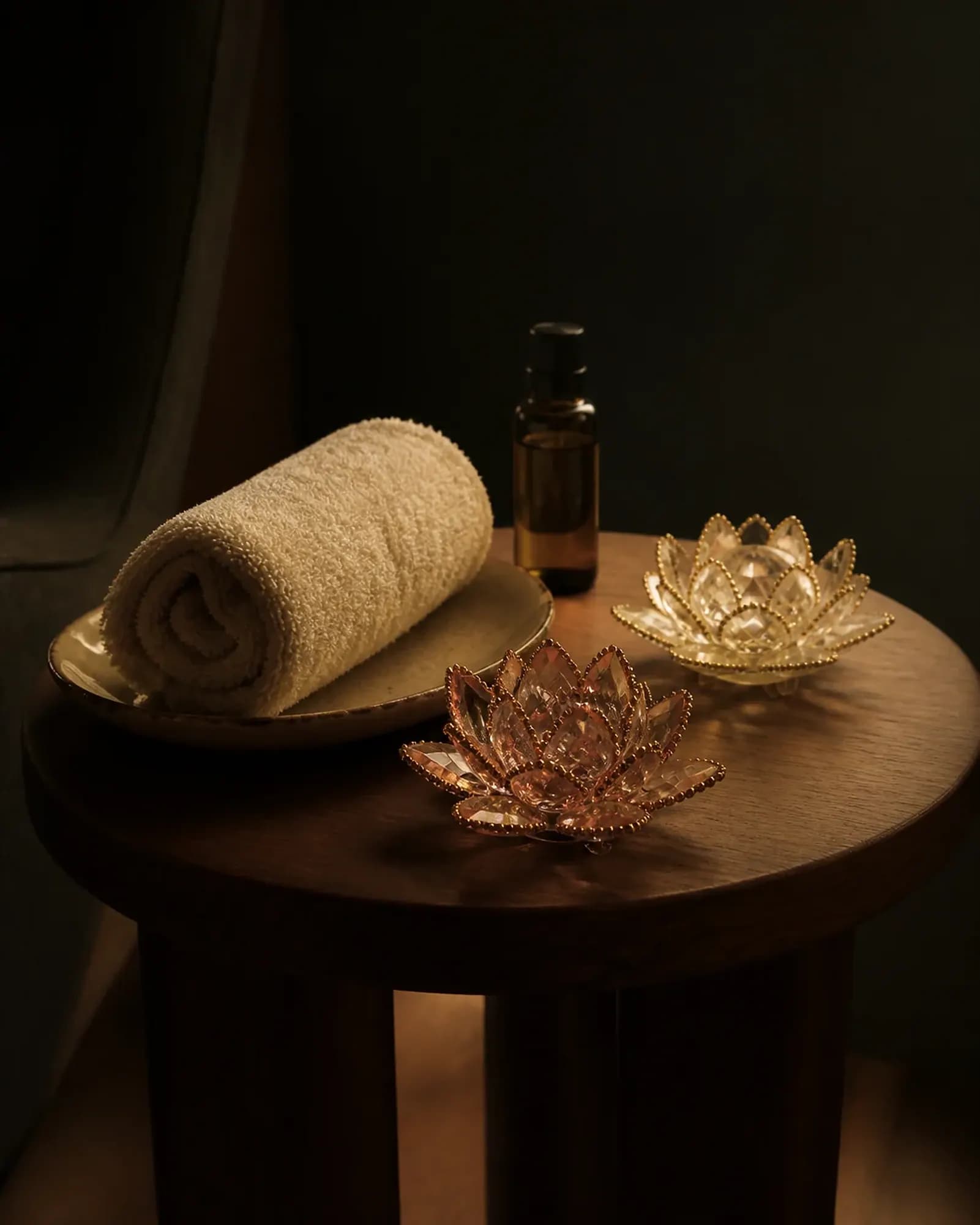 Concept image of a warm towel, amber oil and glass lotus candle holders on dark timber